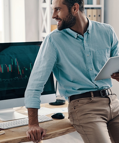 Chico de barba en una oficina sosteniendo una tablet y viendo operaciones en divisas en el ordenador