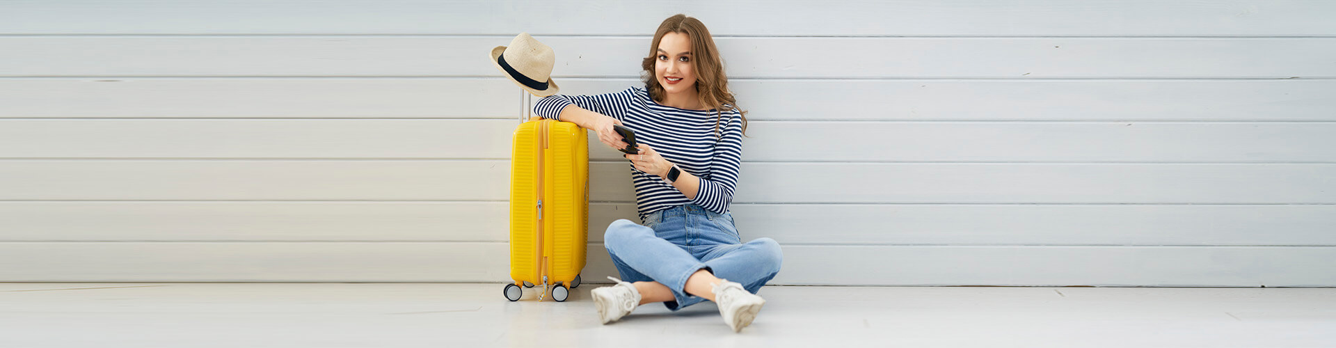 Seguro de Asistencia en Viaje - Mujer joven sentada en el suelo con el movil en la mano, junto a su maleta y un sombrero preparando un viaje Seguro de Asistencia en Viaje - Mujer joven sentada en el suelo con el movil en la mano, junto a su maleta y un sombrero preparando un viaje