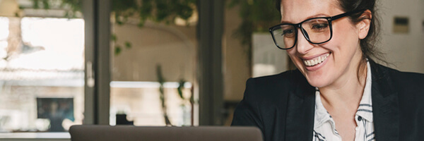 Financiacion a corto plazo / Liquidez - Mujer de negocios morena con gafas y camisa, sonriendo y trabajando frente a su portatil en la oficina Financiacion a corto plazo / Liquidez - Mujer de negocios morena con gafas y camisa, sonriendo y trabajando frente a su portatil en la oficina