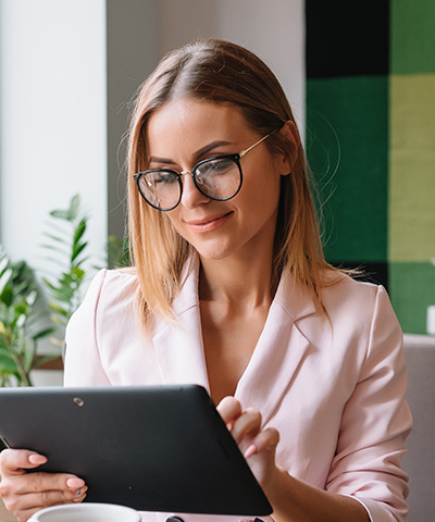 Beneficios de realizar operaciones en divisas  con Globalcaja Beneficios de Operar con Divisas en Globalcaja - Empresaria vestida de traje, con gafas, operando las divisas de su empresa  a través de una Tablet