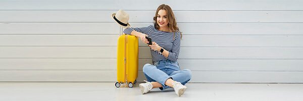 Mujer joven sentada suelo con movil mano junto su maleta y un sombrero preparando viaje - Seguro de viaje Globalcaja