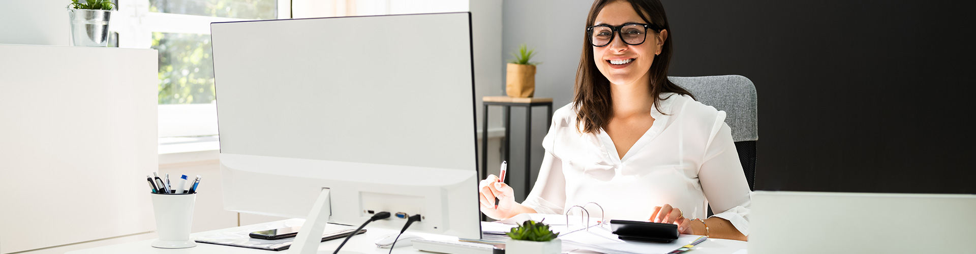 Cuenta de crédito para empresas, financiamiento inmediato de Globalcaja Financiamiento inmediato para empresas a través de Cuenta de crédito - Chica vestida de camisa blanca y gafas negras sujetando un boli, trabajando en la oficina al frente de un ordenador