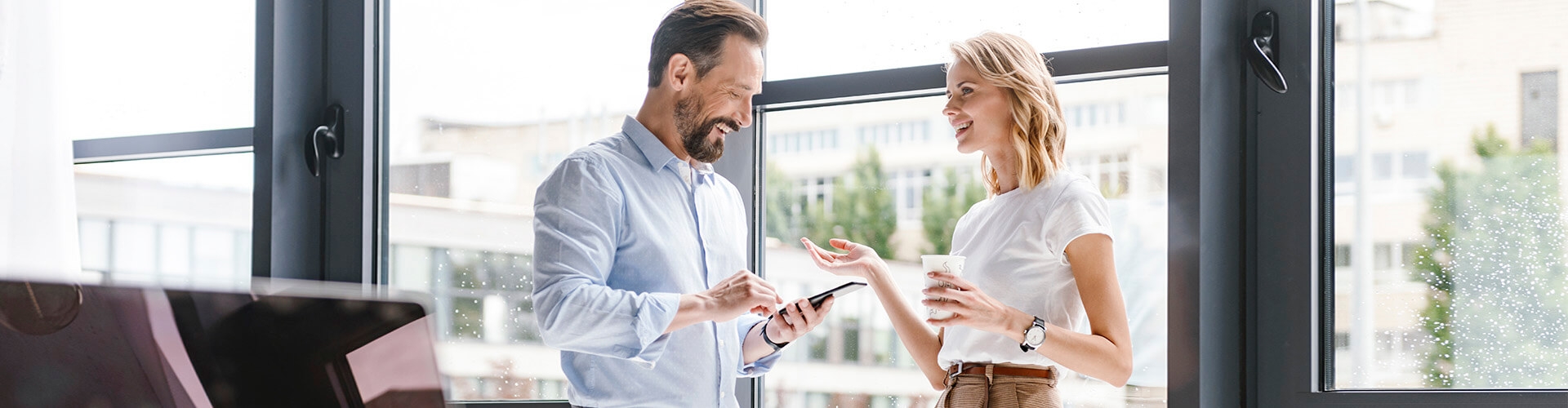 Cartera de Inversión Premium - Hombre con camisa y móvil en la mano, y mujer de negocios con café en la mano sonriendo en la oficina