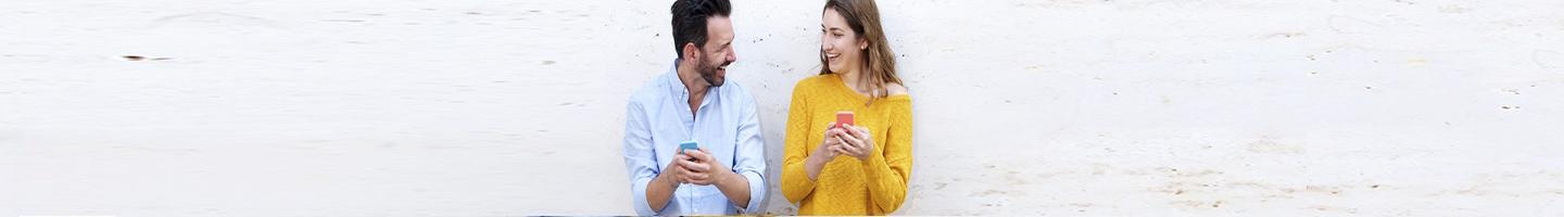 Hazte cliente - Mujer con camiseta amarilla y hombre con camisa azul sonriendo con sus móviles en las manos haciéndose cliente de Globalcaja