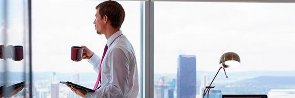 Factoring Internacional - Un hombre con camisa y corbata, con una taza en la mano mirando por la ventana Factoring Internacional - Un hombre con camisa y corbata, con una taza en la mano mirando por la ventana