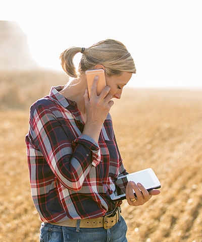 Chica con camisa de cuadros conversando por el móvil y mirando una tablet donde se refleja la producción de los cultivos 